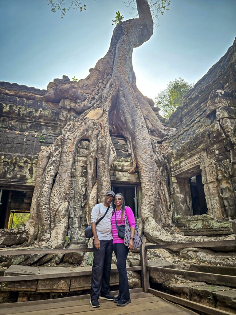 Mike and Marge standing in front of the iconic strangler fig tree engulfing the ancient stone ruins of Ta Prohm temple, Cambodia