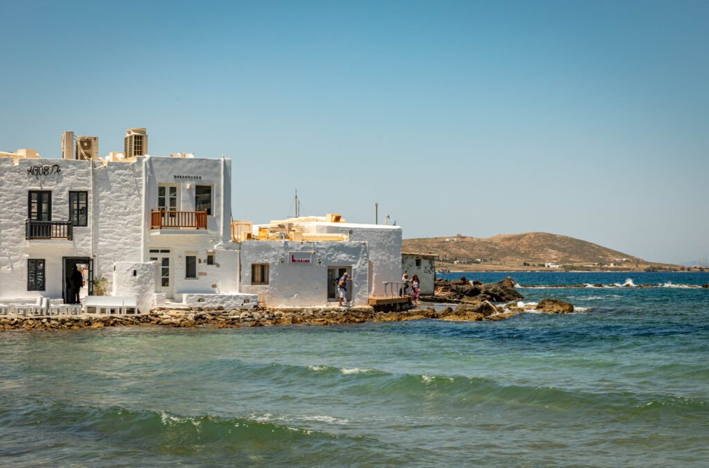 Whitewashed building at the water's edge in Naousa, slow travel Paros Greece — waves breaking against the rocky shoreline